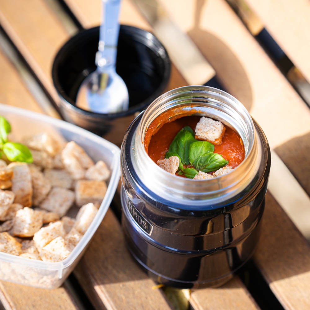 Un thermos aperto rivela una zuppa di pomodoro fresca con basilico e crostini. Accanto al thermos c'è un contenitore con crostini extra e un cucchiaio.