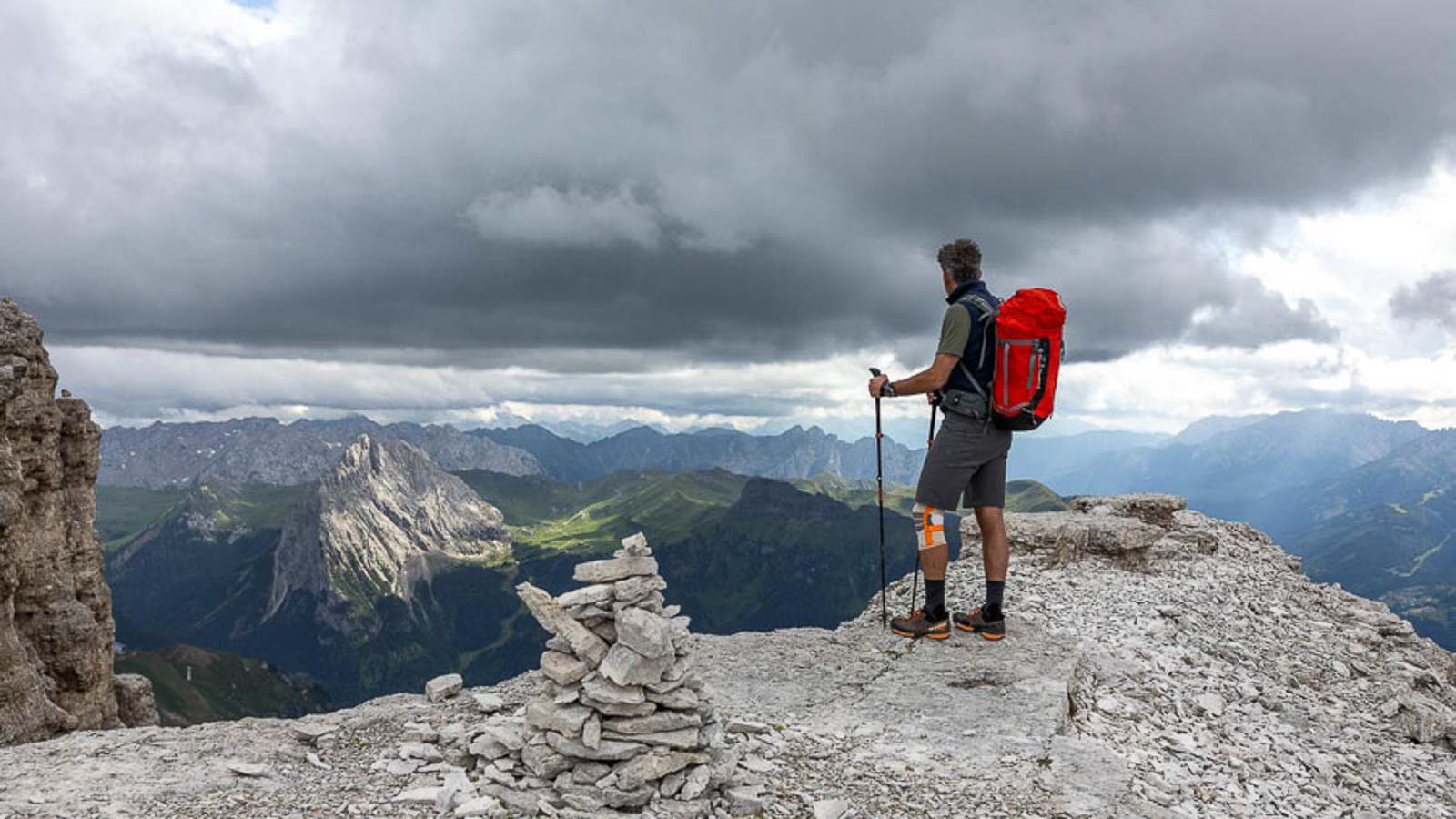 Una persona con uno zaino rosso è in piedi su un affioramento roccioso nelle Dolomiti, guardando lontano. Il cielo è nuvoloso e c'è una costruzione di ometti di pietra in primo piano.