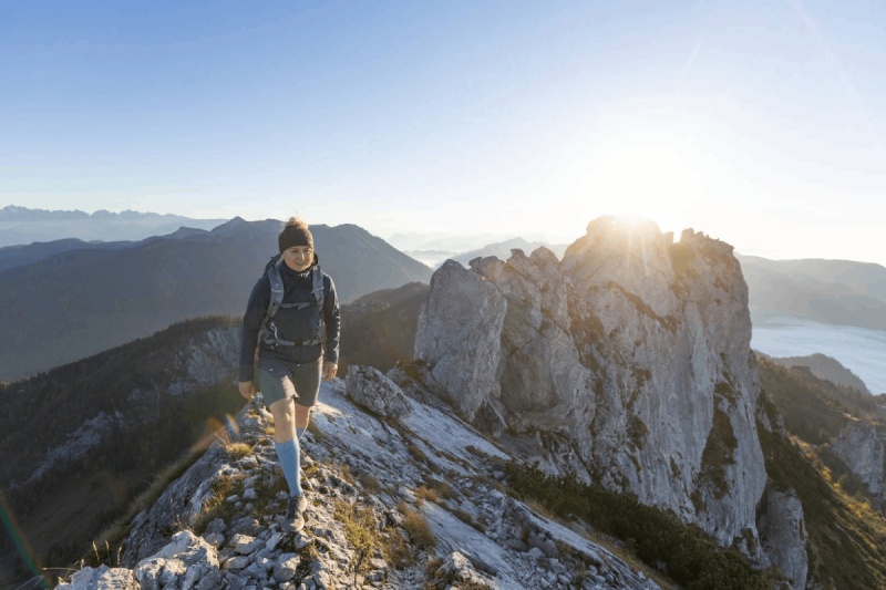 Un'escursionista con Outdoor Merino Compression Socks cammina su un sentiero di montagna all'alba. Questi calzini a compressione offrono supporto ottimale e comfort per escursioni impegnative.