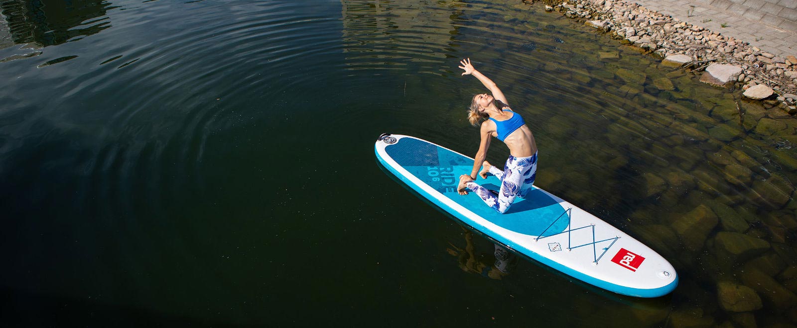 La donna fa l'esercizio di yoga sul tabellone di sup vicino alla riva sott'acqua, alcune pietre sono visibili