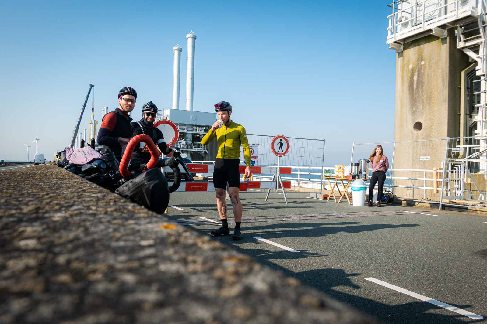 Tre ciclisti aspettano in un cantiere per la costruzione di un ponte, bevendo caffè e mangiando biscotti