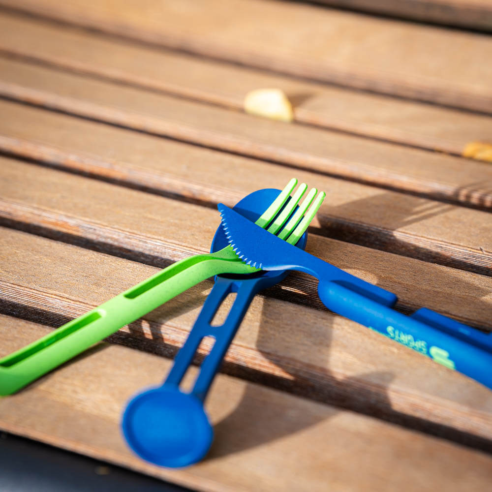 Un set di posate verde e blu è incrociato su una panchina di legno. Il set è ideale per pasti all'aperto e confezioni compatte.