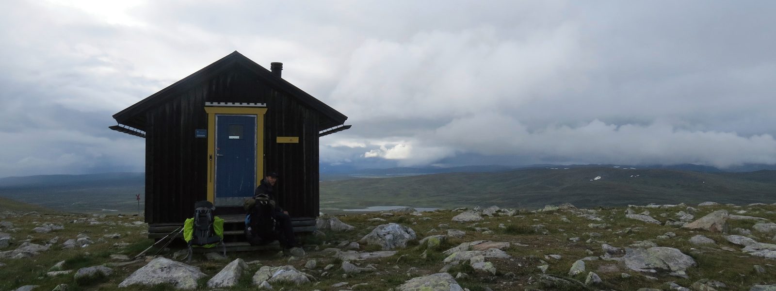 Una capanna solitaria tra le montagne nel mezzo di prati bloccanti con piccole rocce sullo sfondo la fitta copertura nuvolosa e un piccolo lago di montagna