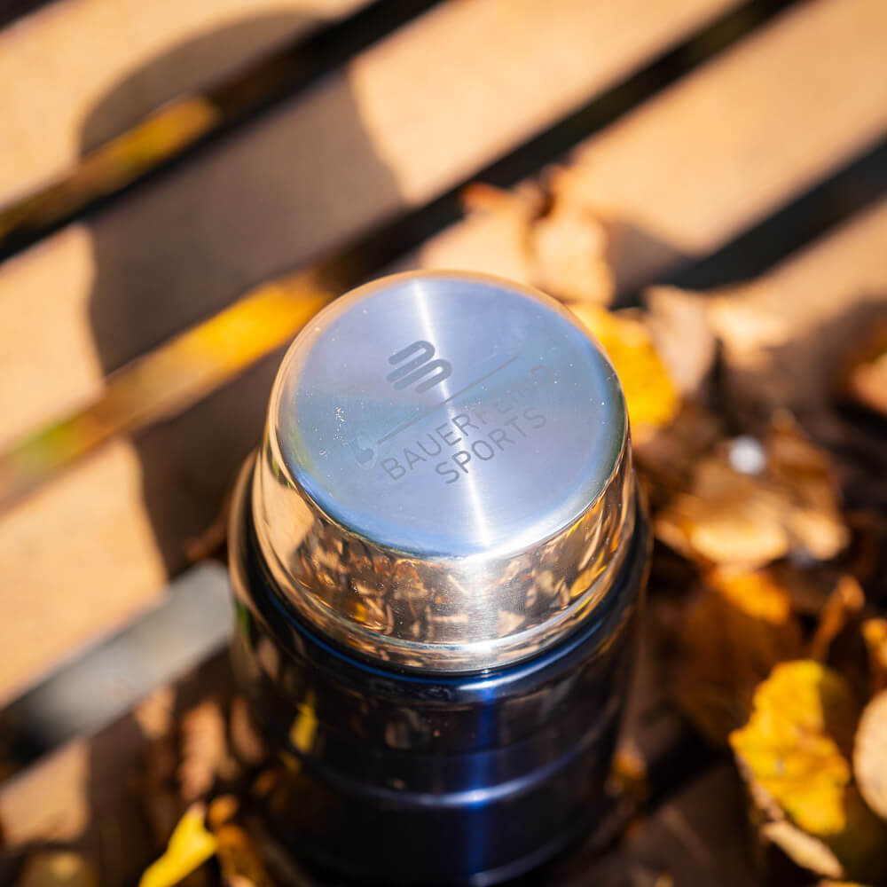 Primo piano del coperchio lucido di un thermos con inciso il logo Bauerfeind Sports. Il coperchio è appoggiato su una panca di legno, circondato da foglie autunnali.