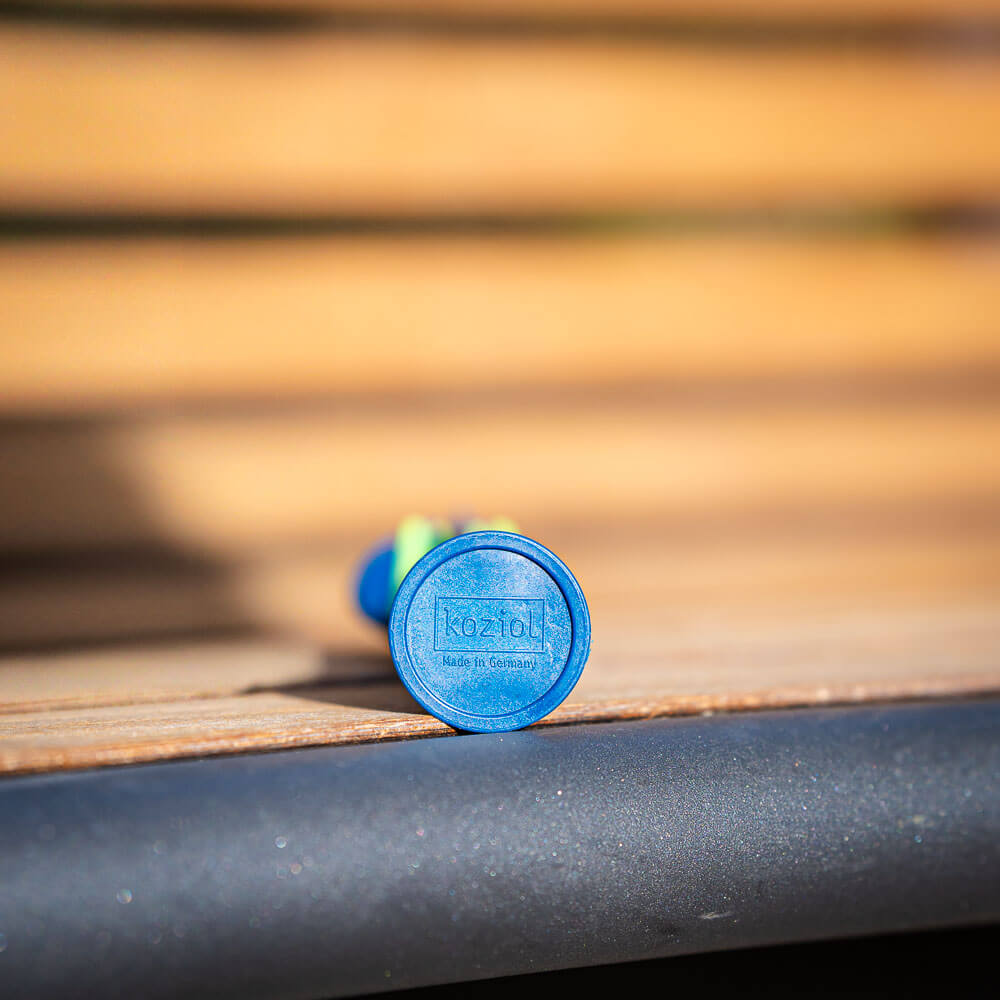 Primo piano del fondo di una posata blu sdraiata su una panchina di legno. Sul fondo è presente il logo "Koziol Made in Germany."