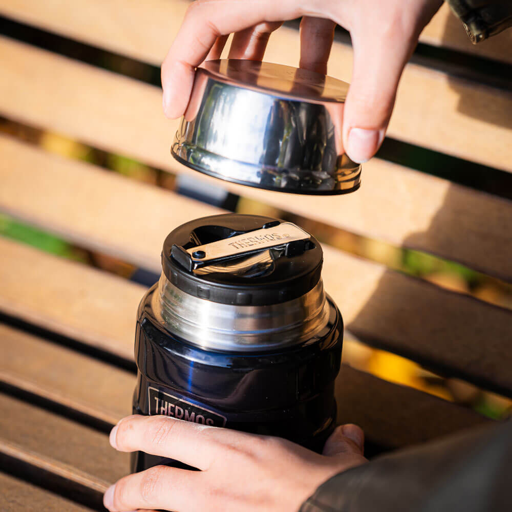 Una mano solleva il coperchio di un thermos per rivelare la chiusura sottostante. Il thermos è appoggiato su una panca di legno in un ambiente autunnale.