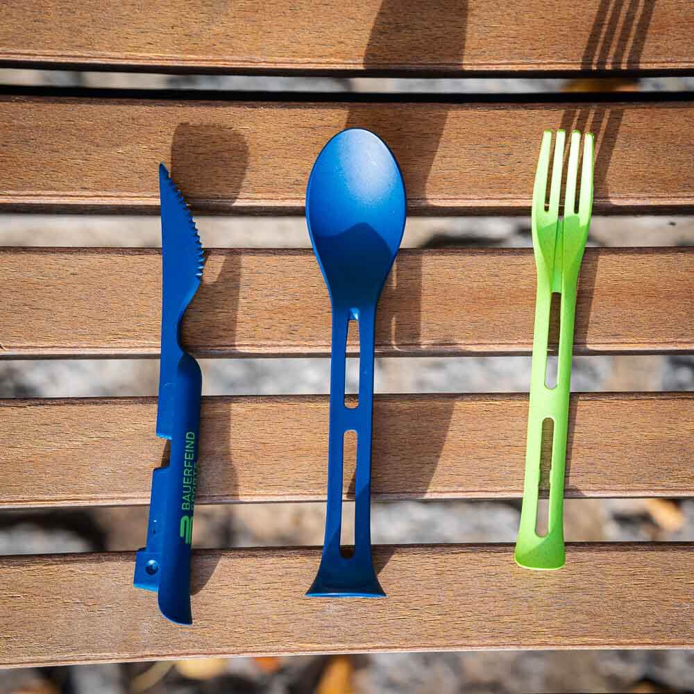 Un set di posate smontato con un coltello blu, un cucchiaio blu e una forchetta verde sono affiancati su una panchina di legno.