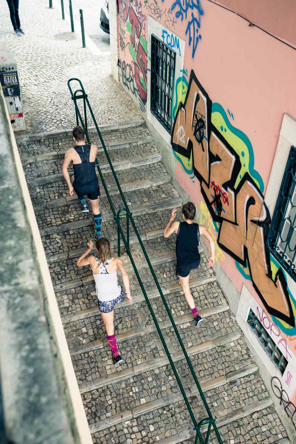 Bird's Eye View: un uomo e due donne in abiti da corsa corrono una scala in una città vecchia con graffiti sui muri